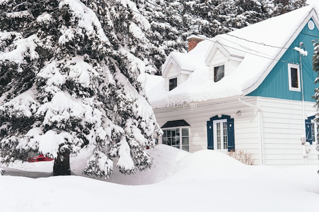 A snow-covered house surrounded by deep drifts and frosted evergreen trees, illustrating why understanding how to winterize plumbing is essential during harsh winter conditions.